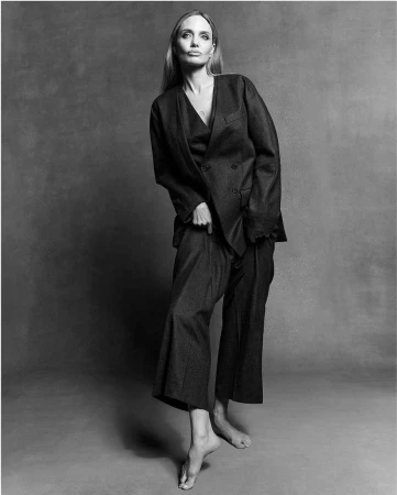Black and white portrait of Angelina Jolie wearing a dark oversized suit against a textured grey background.