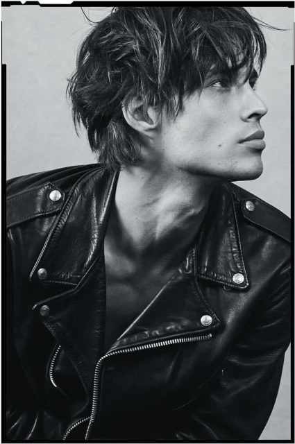 Monochrome portrait of a fair-skinned adult male fashion model wearing a black leather jacket, looking off-camera in a studio setting.