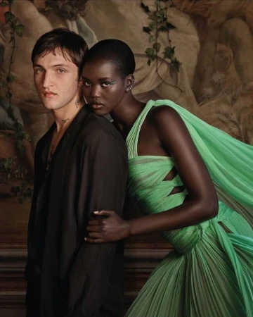 A fashion editorial featuring a caucasian man and a black woman wearing a pastel green gown, posing in front of a Renaissance-style backdrop.