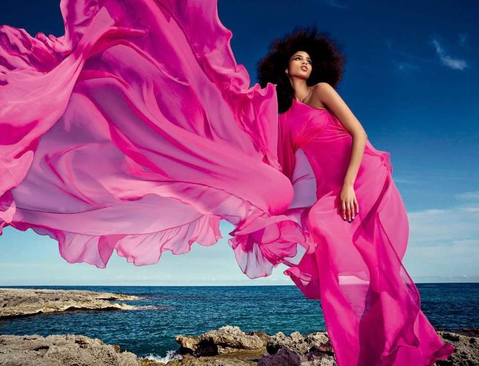 Woman in a flowing pink dress poses dramatically by the ocean under a blue sky, creating a striking fashion and nature image.