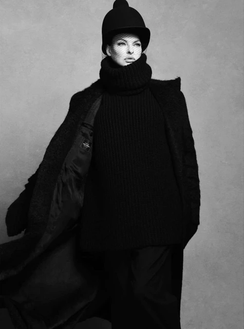 Monochrome fashion portrait of woman in black turtleneck, coat, and riding helmet with net veil against a muted gray backdrop.