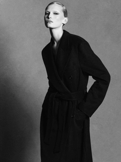 A slender woman poses in a long, belted black coat against a gray backdrop, showcasing minimal makeup and a sleek, pulled-back hairstyle.