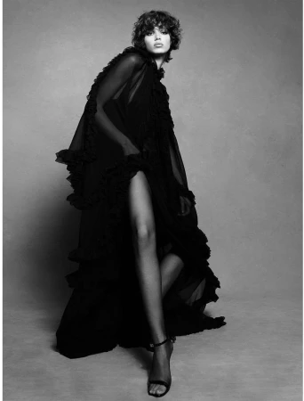 Model with short curly hair wearing a long black ruffled sheer gown against a plain grey studio backdrop.