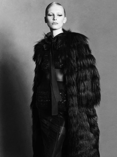 Black and white fashion portrait of a slender adult female wearing a long fur coat and posing in a studio setting. 