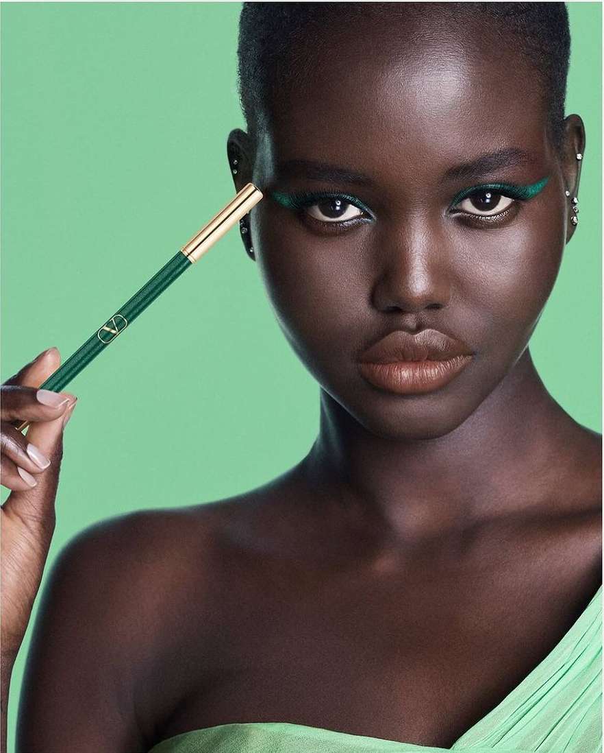 Close-up of a dark-skinned woman with green eyeliner, holding a green eyeliner brush against a light green background.