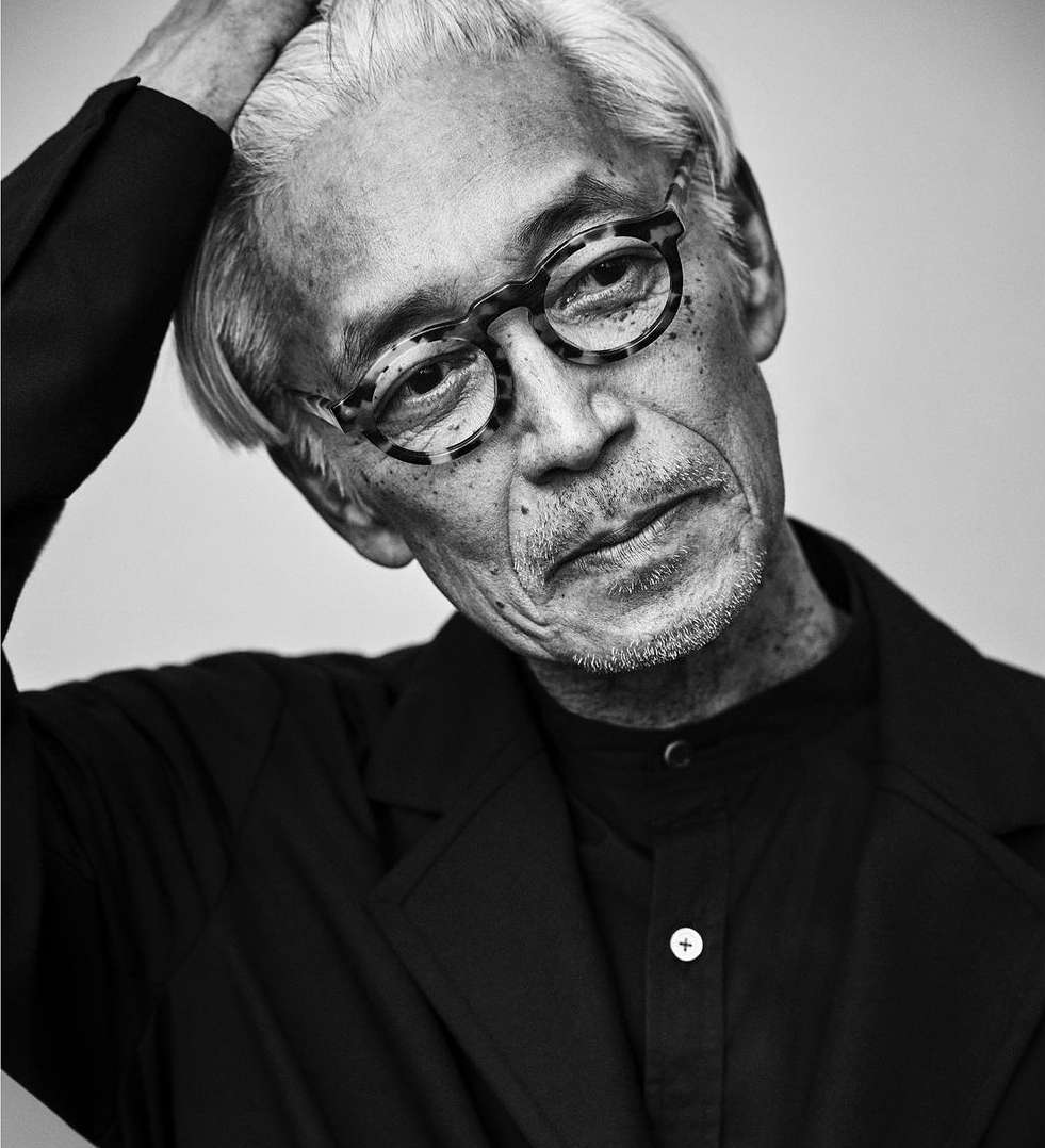 Black and white portrait of an elderly adult male with light gray hair wearing tortoise-shell glasses and a dark blazer in a studio.
