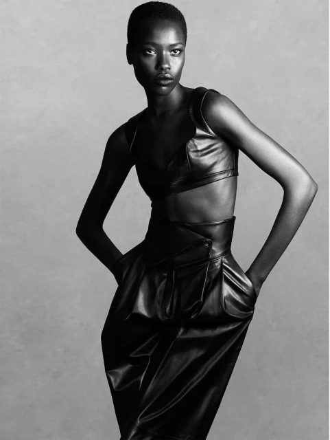 A striking studio shot of a Black woman in a stylish black leather outfit, standing with hands in pockets against a neutral background.