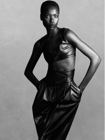 A striking studio shot of a Black woman in a stylish black leather outfit, standing with hands in pockets against a neutral background.