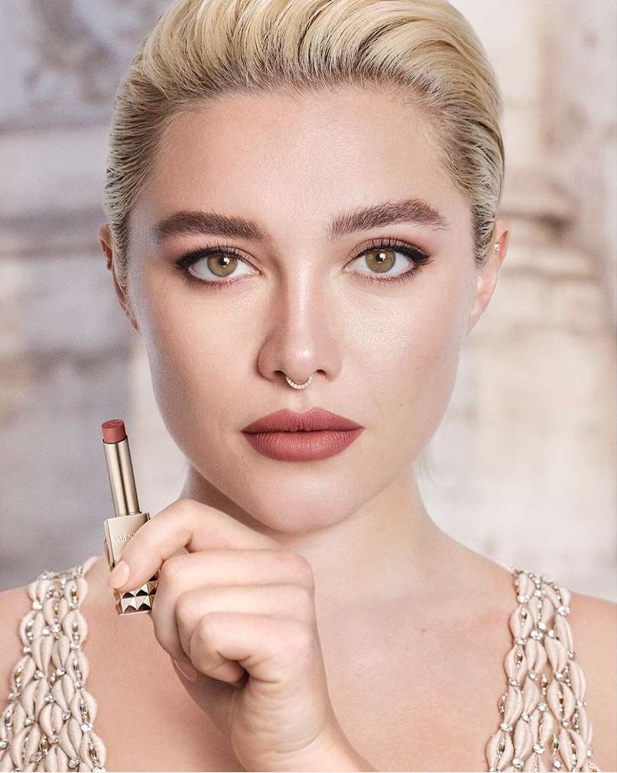 Close-up portrait of Florence Pugh holding a lipstick tube, featuring blonde hair, septum ring, and embellished dress.