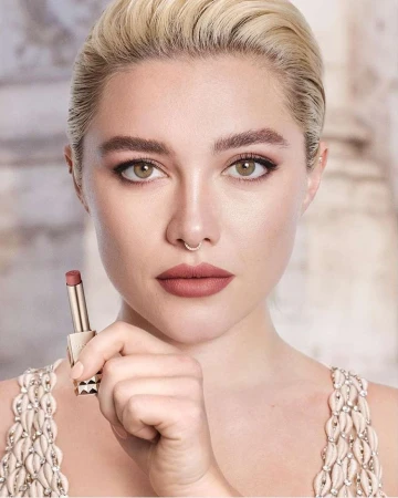 Close-up portrait of Florence Pugh holding a lipstick tube, featuring blonde hair, septum ring, and embellished dress.