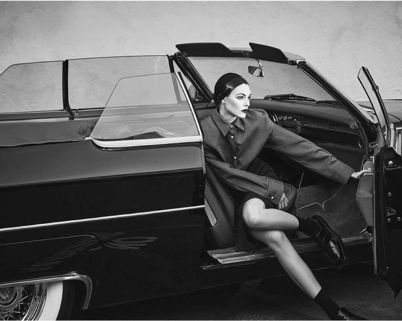 Monochrome image of a fashion model posing in the doorway of a classic black convertible car with an open door and light grey background.