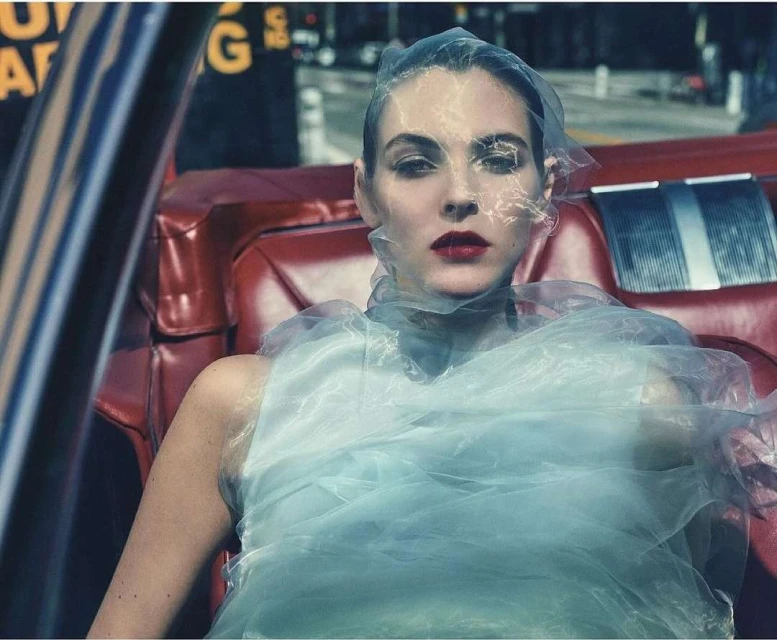 Woman in red car wearing translucent plastic material as clothing. Natural lighting in outdoor urban setting. Beauty, fashion.