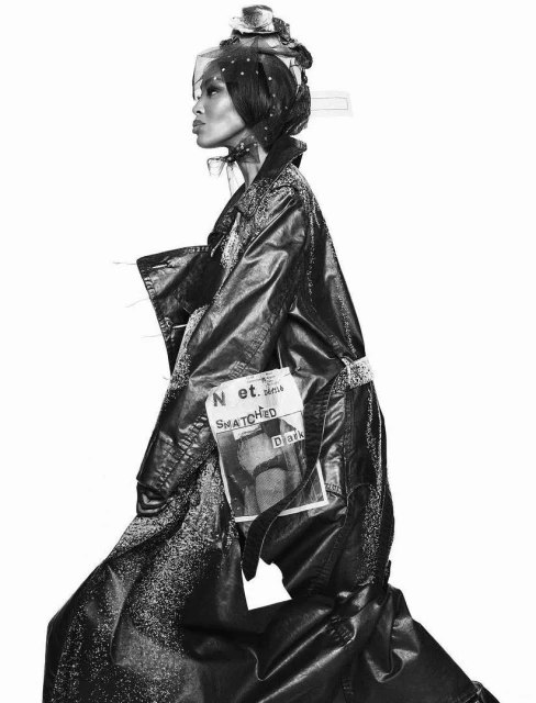 Black and white fashion portrait of a woman in a textured black coat and headscarf against a white background in a studio setting.