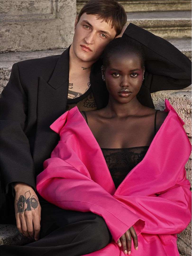 Fashion editorial shot of a young couple posing on stone steps. Woman wears a pink top, man wears a black jacket, set against a stone background.