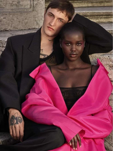 Fashion editorial shot of a young couple posing on stone steps. Woman wears a pink top, man wears a black jacket, set against a stone background.