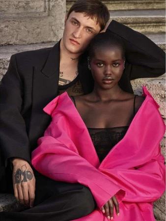 Fashion editorial shot of a young couple posing on stone steps. Woman wears a pink top, man wears a black jacket, set against a stone background.