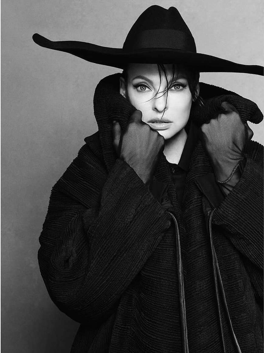 Black and white portrait of a woman wearing a wide-brimmed hat and textured coat, set against a neutral grey backdrop in a photo studio.