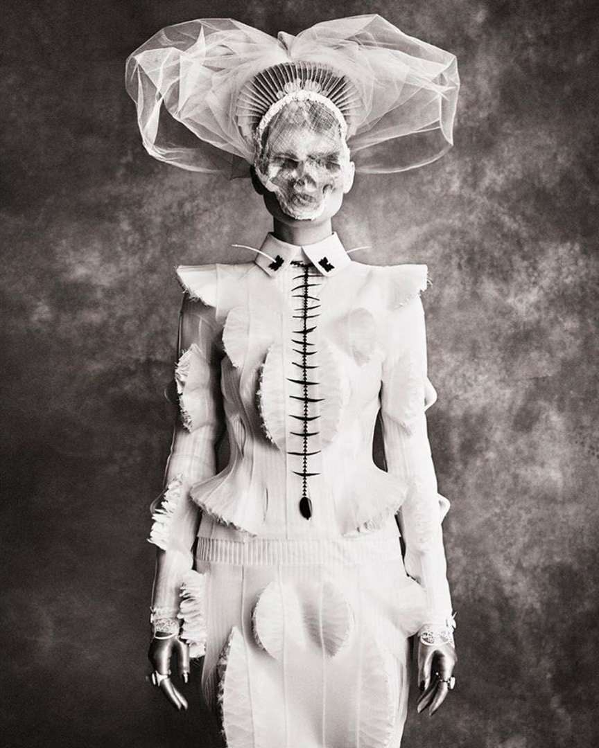 Monochrome portrait of person in a white couture outfit and elaborate headpiece with a skull face in front of a mottled backdrop.