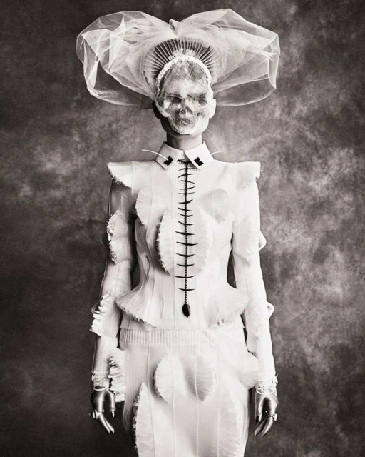 Monochrome portrait of person in a white couture outfit and elaborate headpiece with a skull face in front of a mottled backdrop.