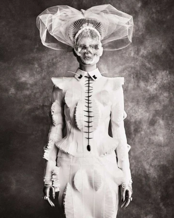 Monochrome portrait of person in a white couture outfit and elaborate headpiece with a skull face in front of a mottled backdrop.
