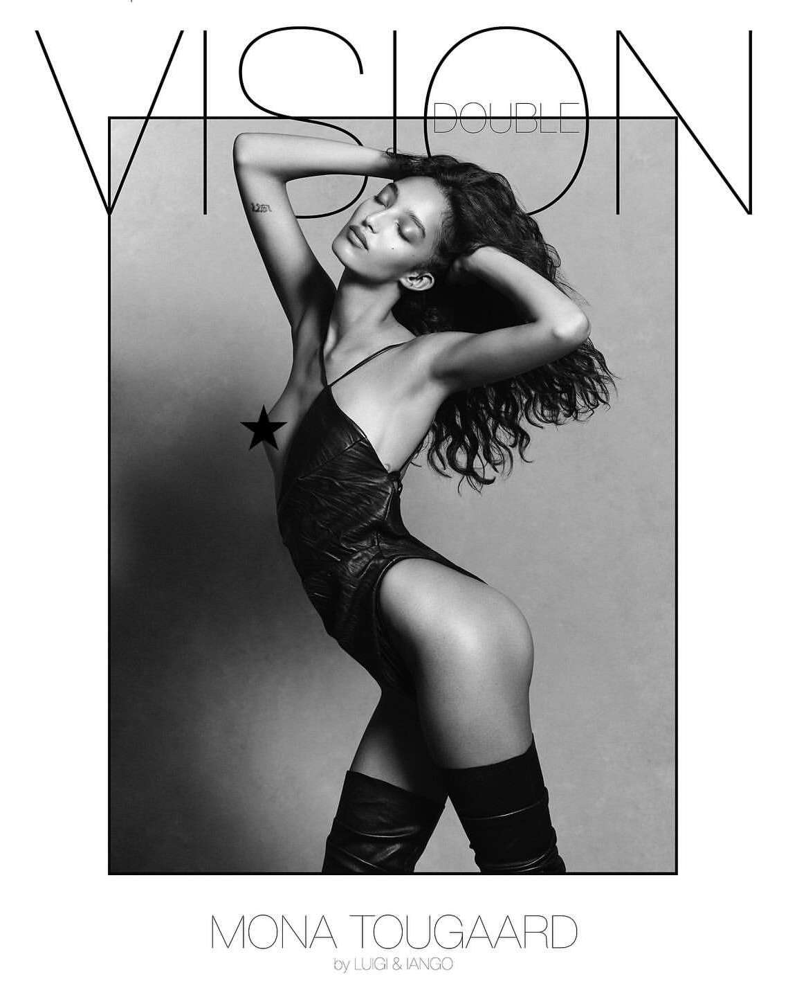 A woman with long curly hair poses in a black bodysuit and black boots against a grayscale backdrop for a studio fashion shot.