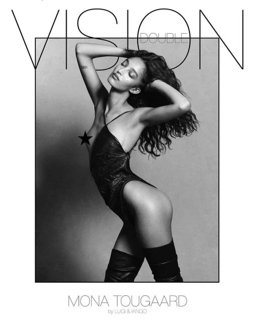 A woman with long curly hair poses in a black bodysuit and black boots against a grayscale backdrop for a studio fashion shot.