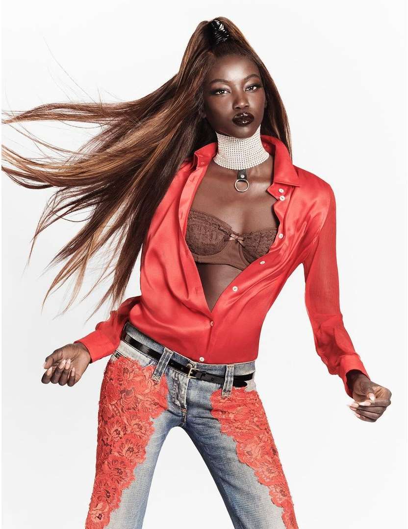 An African American fashion model poses in an unbuttoned red blouse, blue jeans with red lace, and a pearl choker in a studio shot.