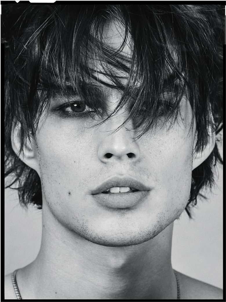 Monochrome portrait of a young adult male with dark hair, eye makeup, and a serious expression. Visible necklace on a neutral background.