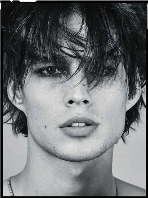 Monochrome portrait of a young adult male with dark hair, eye makeup, and a serious expression. Visible necklace on a neutral background.