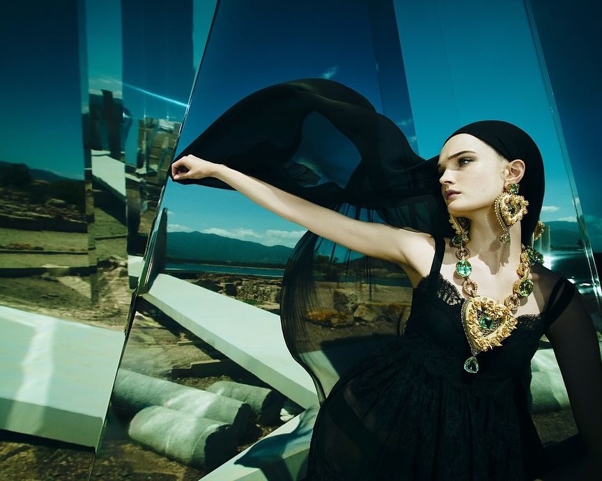 Adult fashion model wearing a black dress and jewelry posing in a mirrored environment with a landscape background and bright lighting.