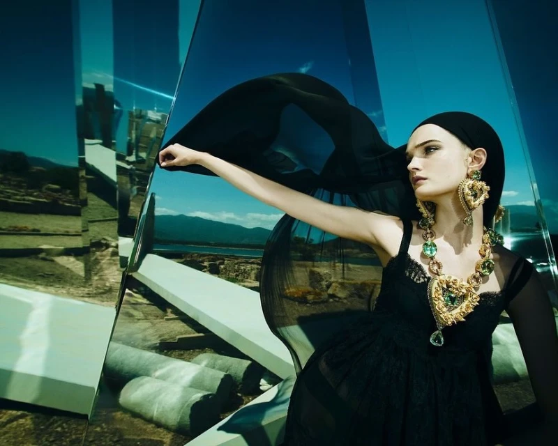 Adult fashion model wearing a black dress and jewelry posing in a mirrored environment with a landscape background and bright lighting.