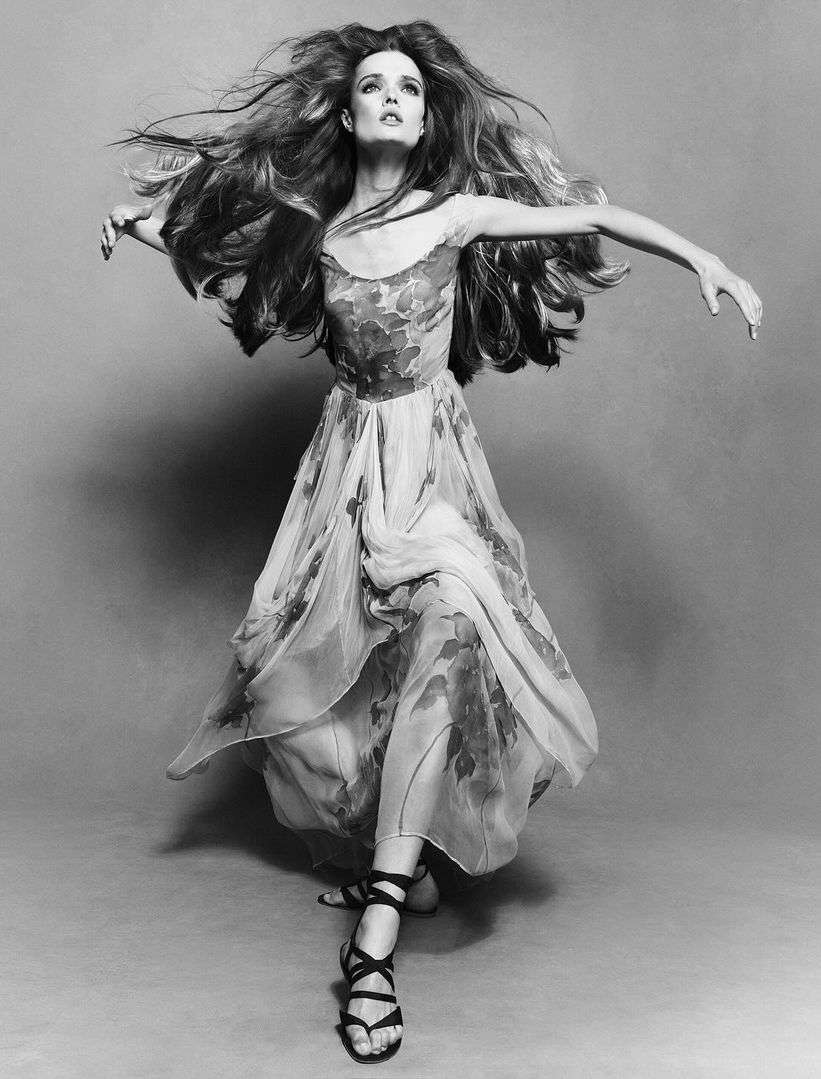 Black and white portrait of a female model with flowing hair wearing a floral dress and strappy sandals against a grey backdrop