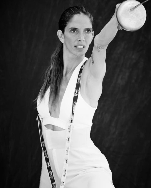 Black-and-white shot of a woman in a white outfit holding a fencing foil, her arm raised, against a dark background.