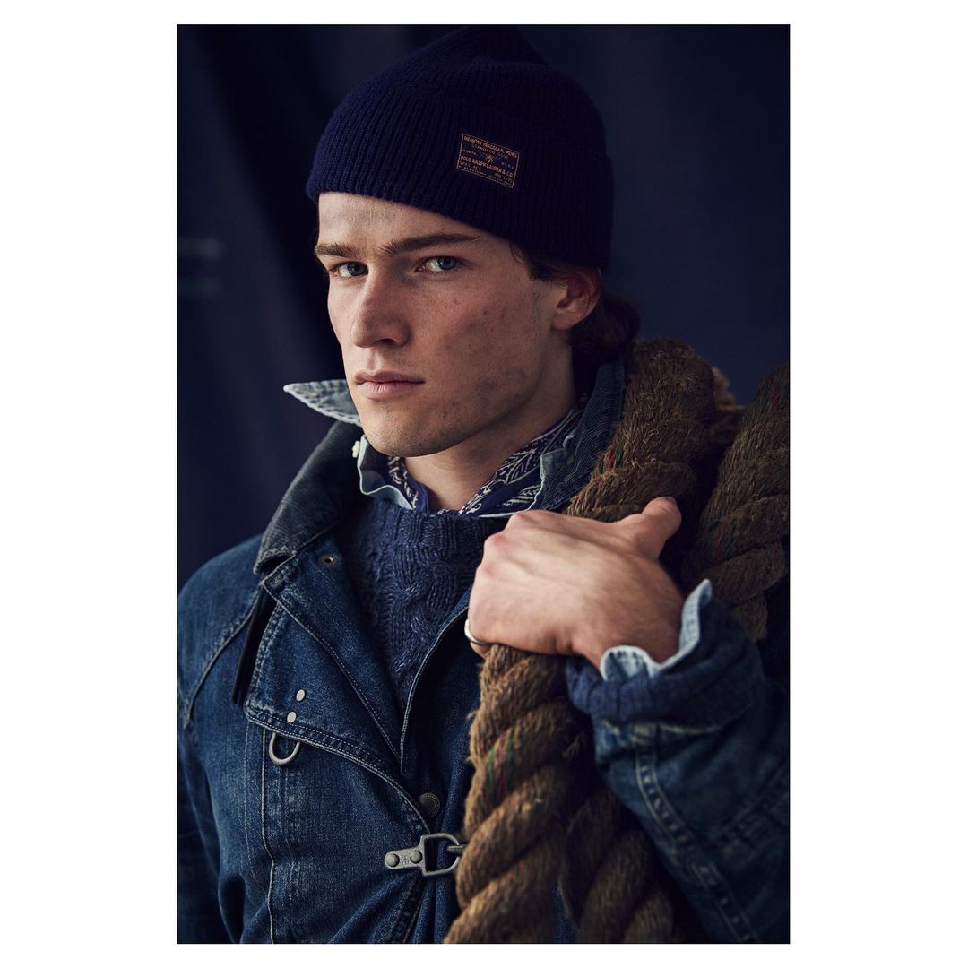 Close-up portrait of a man wearing a dark beanie and denim jacket carrying thick rope over his shoulder.
