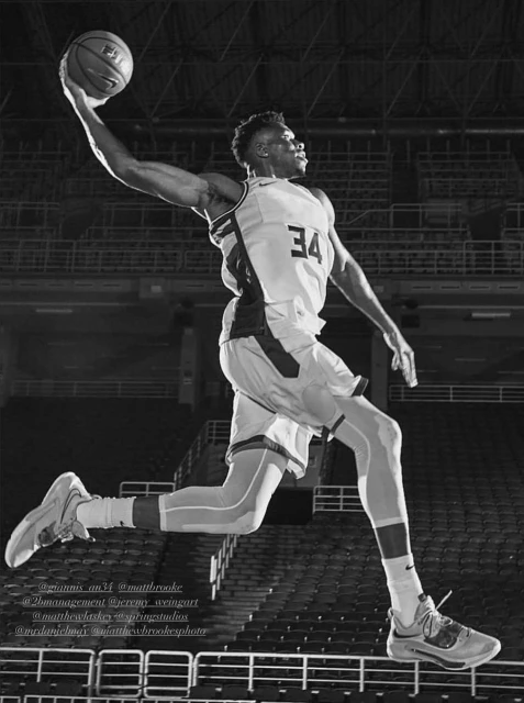 Black and white shot of a basketball player with ball in mid-air, wearing jersey number 34 on an indoor court with stadium seating.