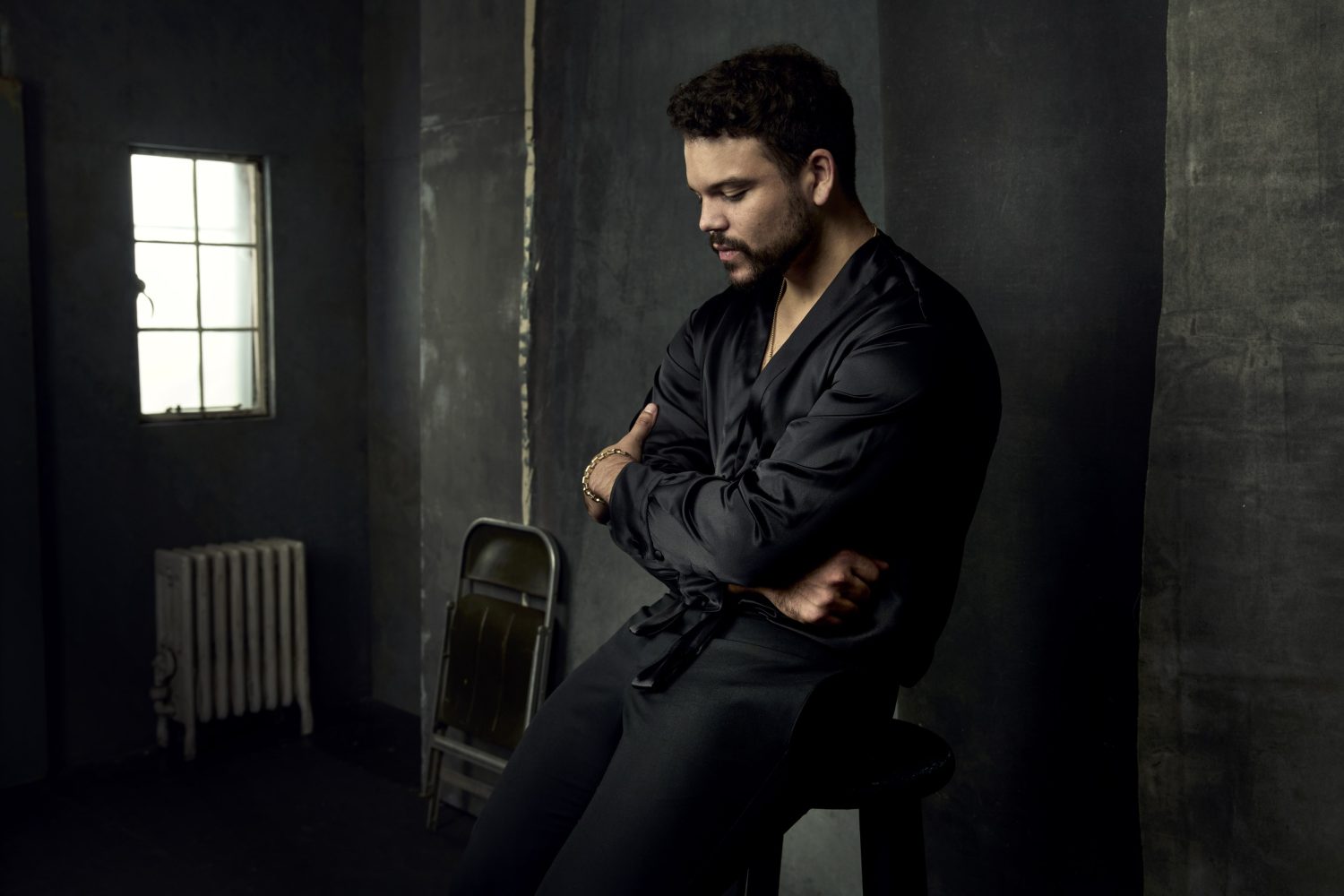 Portrait of a man in a dark robe sitting on a stool in a dim room with a window and radiator, arms crossed and head tilted down.