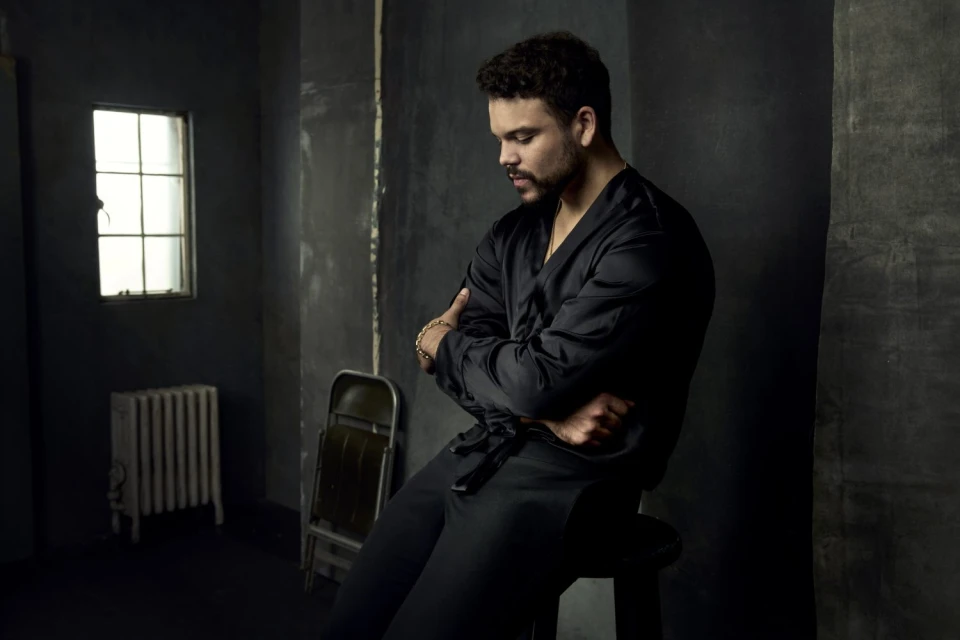 Portrait of a man in a dark robe sitting on a stool in a dim room with a window and radiator, arms crossed and head tilted down.