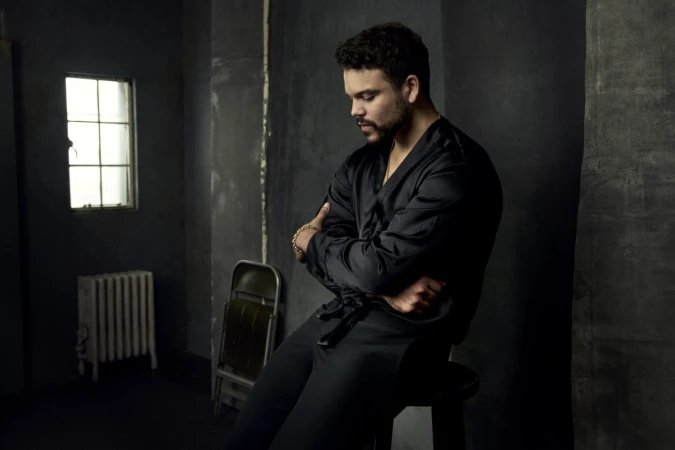 Portrait of a man in a dark robe sitting on a stool in a dim room with a window and radiator, arms crossed and head tilted down.