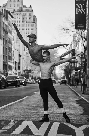 Two shirtless ballet dancers pose en pointe in a New York City street, one carrying the other, with buildings and cars in the background.