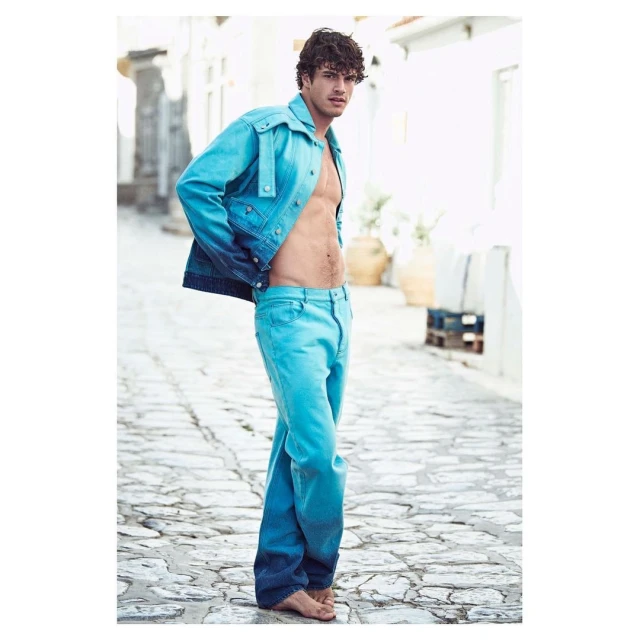 Male fashion model poses in a blue ombre denim jacket and matching denim pants on a cobblestone street, with white buildings in the background.
