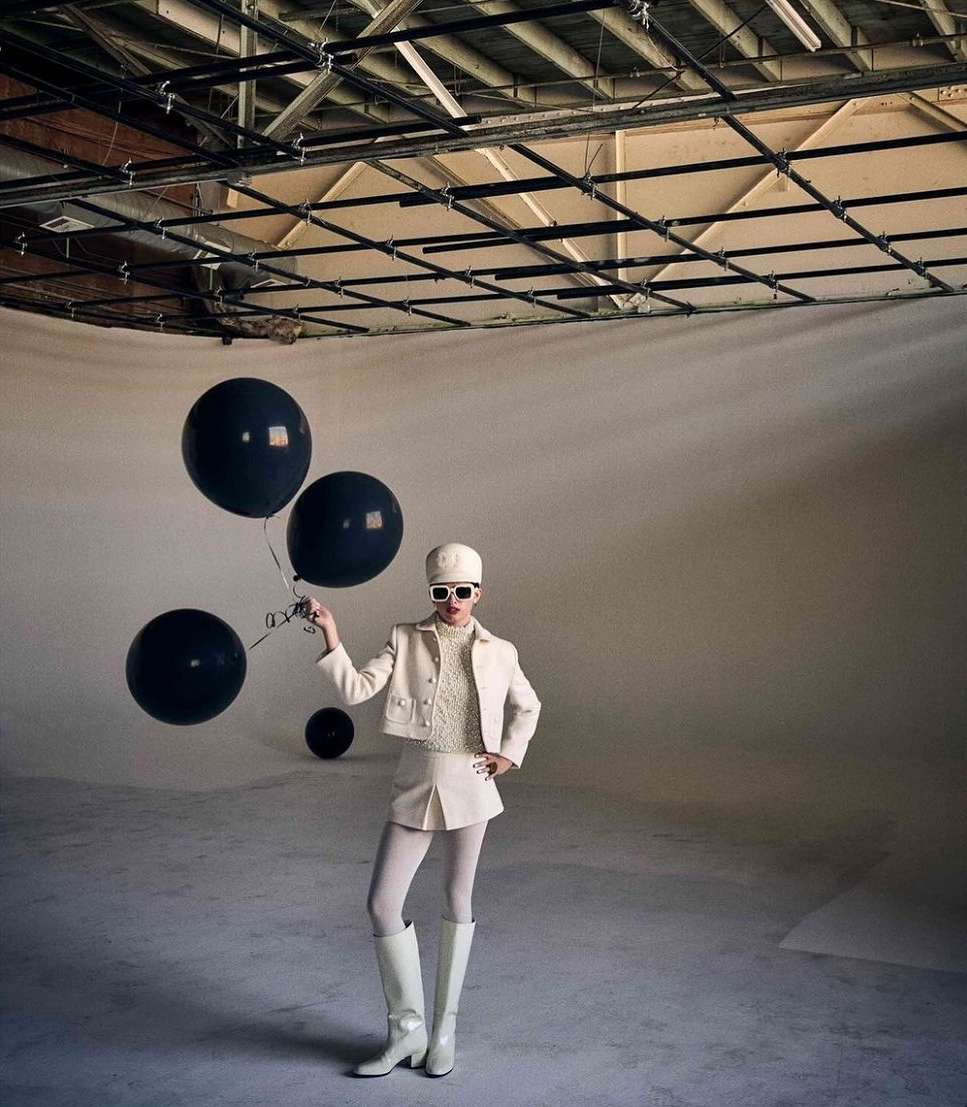 Woman poses in a studio wearing a white outfit, knee-high boots, a hat, and sunglasses, holding black balloons against a neutral backdrop.