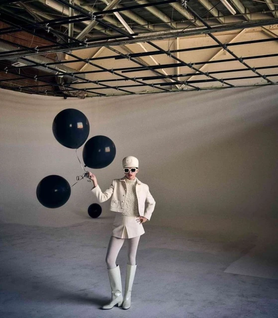Woman poses in a studio wearing a white outfit, knee-high boots, a hat, and sunglasses, holding black balloons against a neutral backdrop.