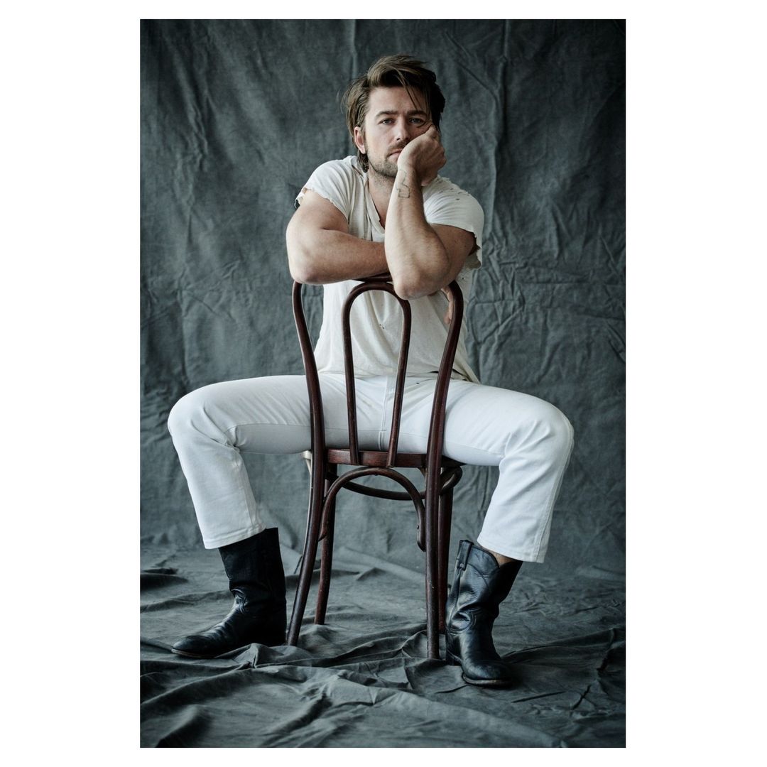 Portrait of a man sitting backwards on a chair in white jeans, t-shirt, and black boots against a textured grey backdrop.