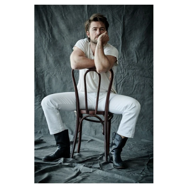 Portrait of a man sitting backwards on a chair in white jeans, t-shirt, and black boots against a textured grey backdrop.