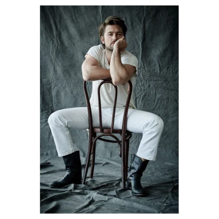 Portrait of a man sitting backwards on a chair in white jeans, t-shirt, and black boots against a textured grey backdrop.