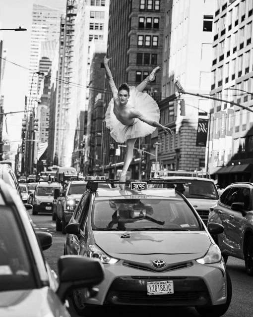 Black and white street photography captures a ballerina in tutu en pointe on a yellow taxi cab amidst New York City traffic and skyscrapers.