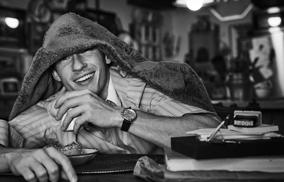 Black and white portrait of a smiling man indoors with a watch, covered by a piece of fabric, seated at a table with food and books.