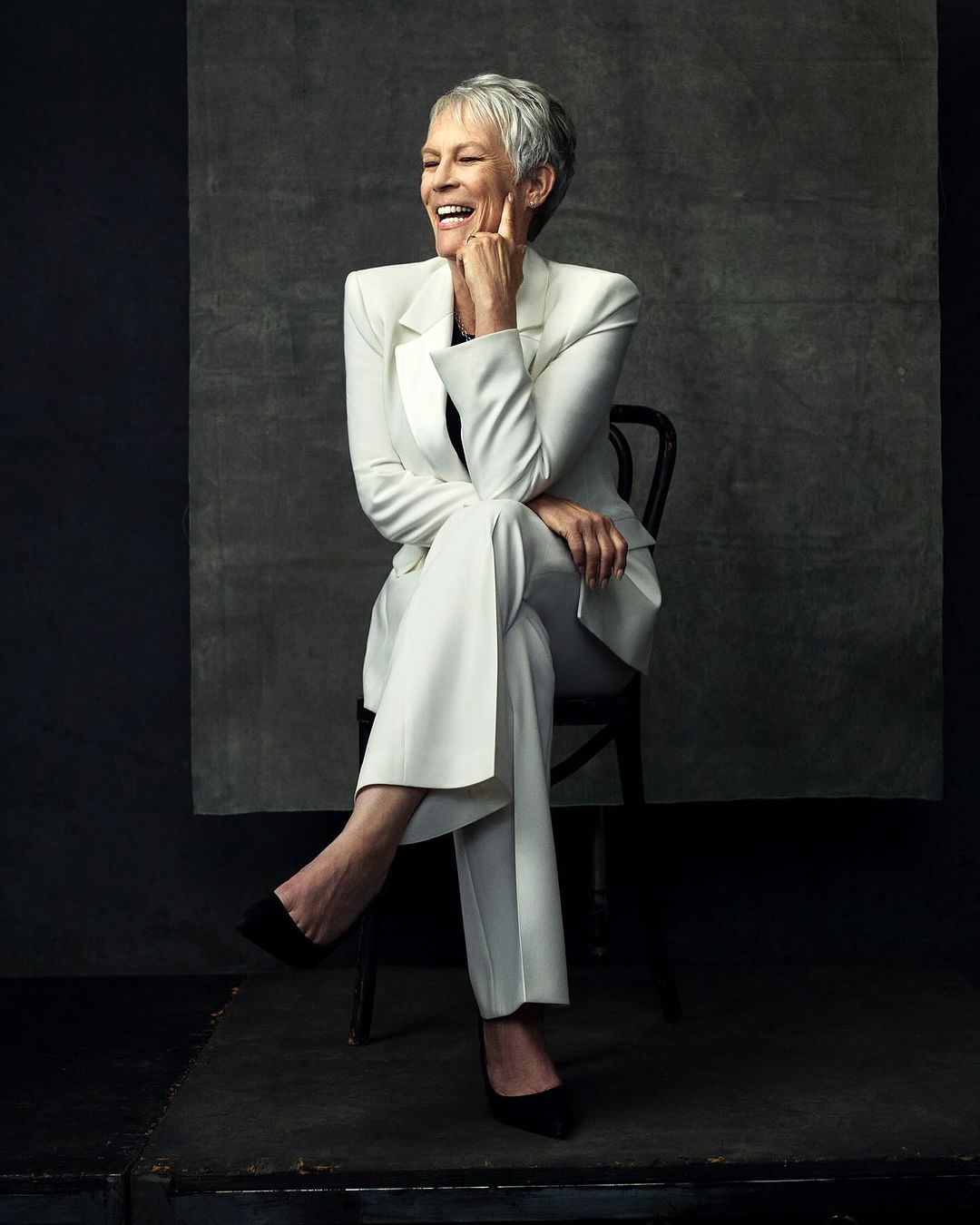 Mature woman with short gray hair smiles while seated in a black chair, wearing a tailored white pantsuit and black heels against gray backdrop.