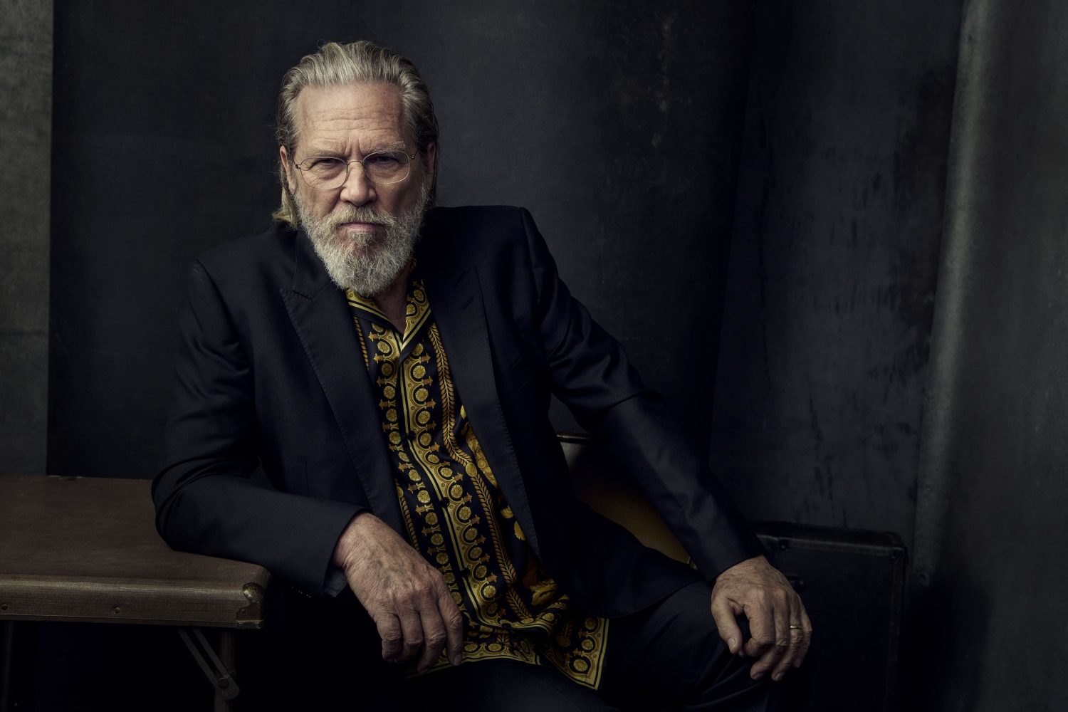 Studio shot of Jeff Bridges wearing a black suit and patterned shirt, seated with a serious expression against a dark textured backdrop.