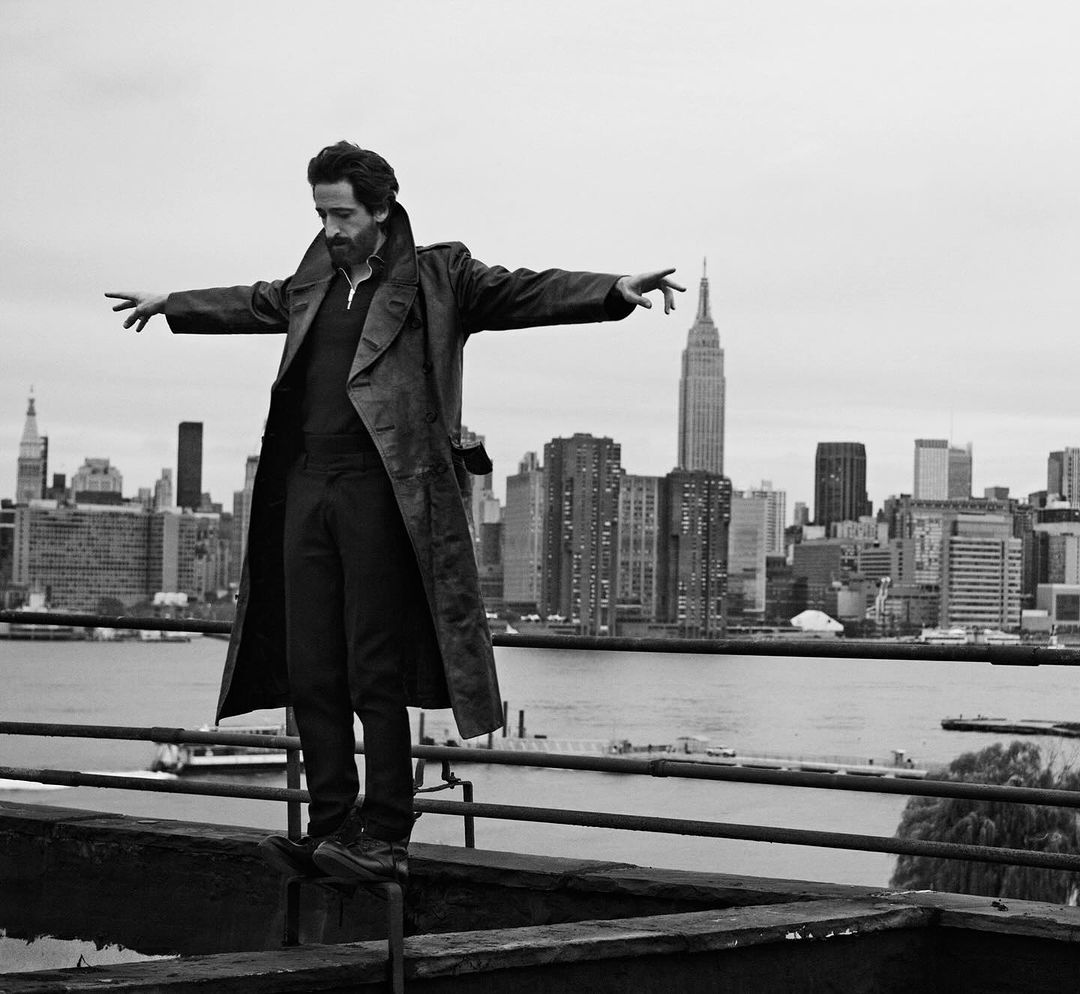 Black and white shot of a man in a trench coat standing on a rooftop ledge with arms outstretched, set against the New York City skyline.